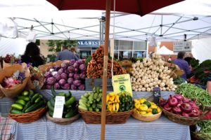 A table adorned with various vegetables, sheltered by a colorful umbrella, set against the backdrop of Capitola, California.