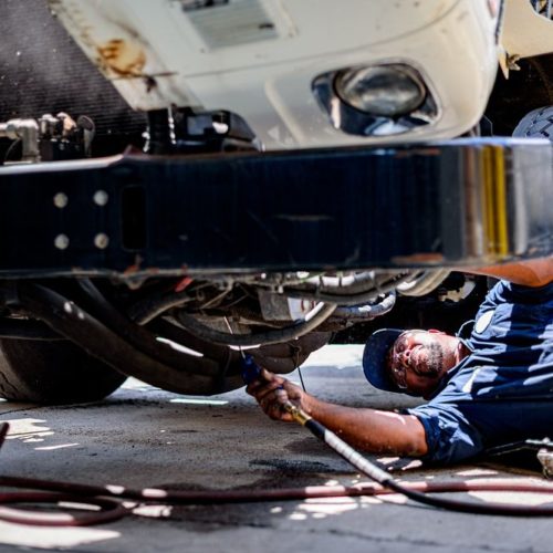 A man is lying on the ground, performing a commercial truck engine repair underneath the vehicle.