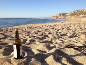 A bottle and glass of wine placed on the sandy beach of Santa Cruz, California, with the serene ocean in the background.